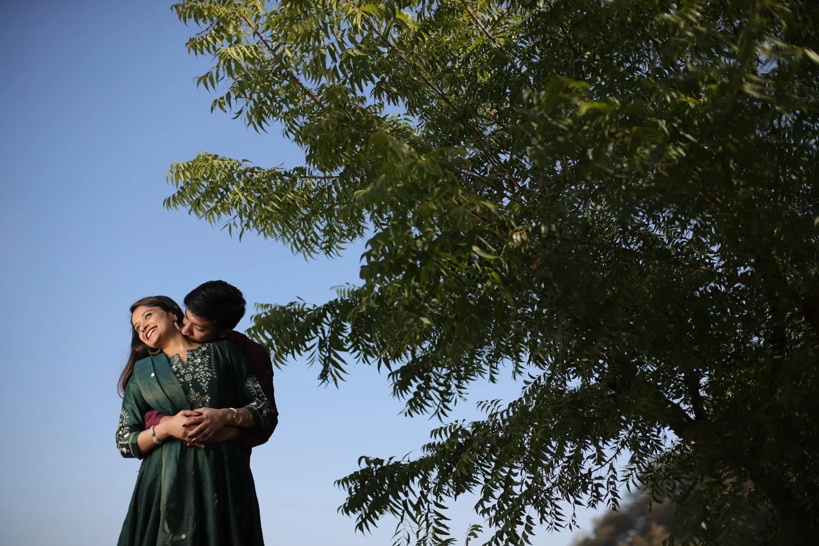 Couple embracing under a neem tree against a clear blue sky during a pre-wedding shoot